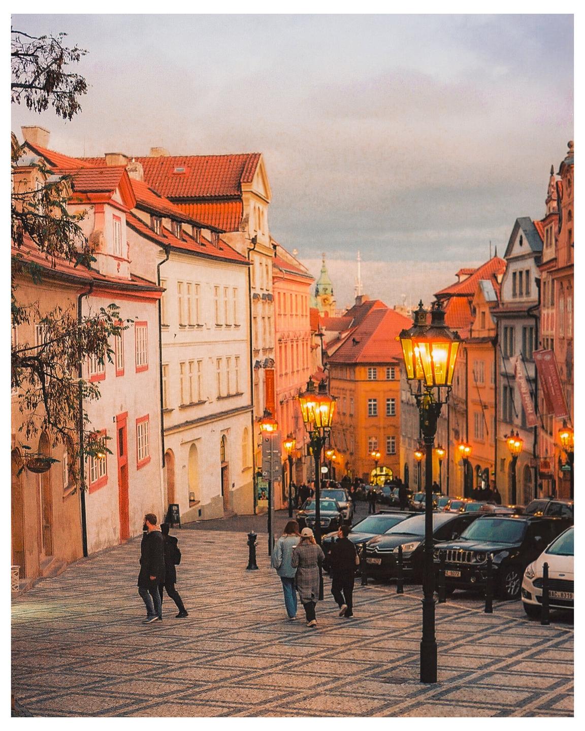 People walk on a cobblestone street lined with buildings and streetlights at dusk.