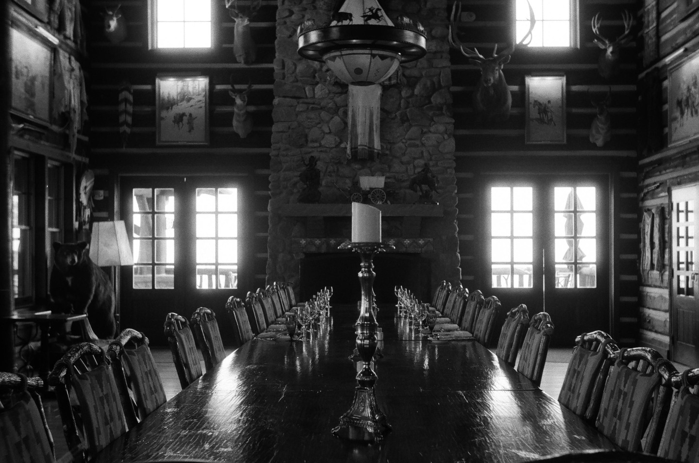 A long dining table with chairs is set in a rustic room with a stone fireplace, taxidermy, and bright windows.
