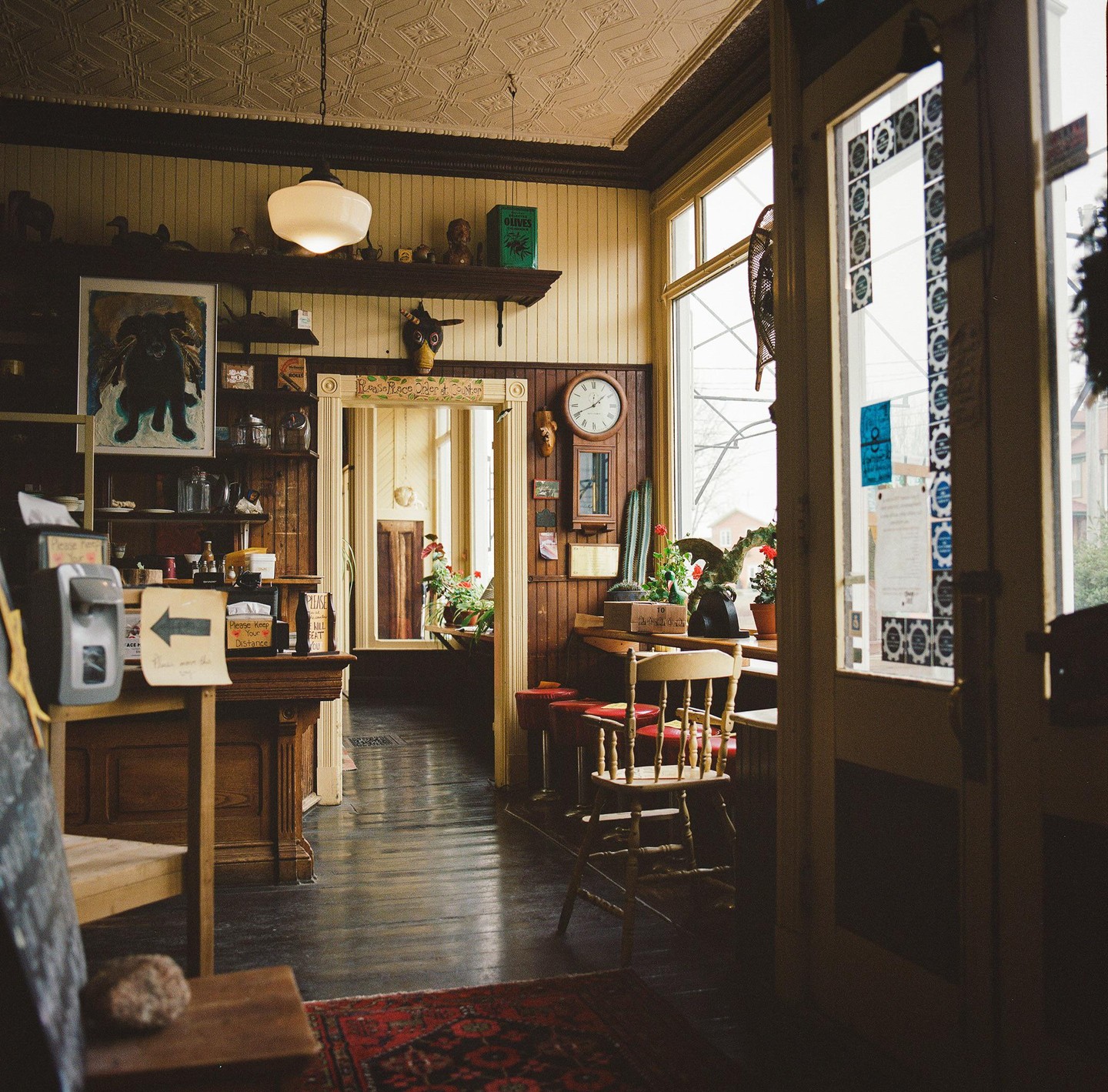 A cozy bakery interior features wooden furniture, potted plants, and a large window looking out onto a street.
