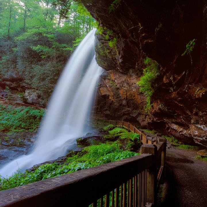 A long exposure photograph captures a waterfall cascading over a rocky cliff face, with a wooden walkway and lush green foliage in the foreground.