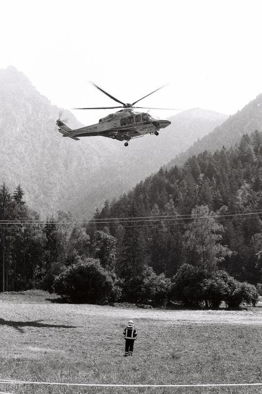 A helicopter takes off over a field where a person in uniform stands, with mountains and trees in the background.