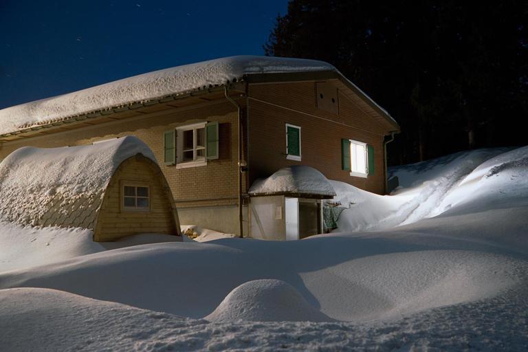 A house with snow on its roof and surrounding ground is illuminated by a single window at night.