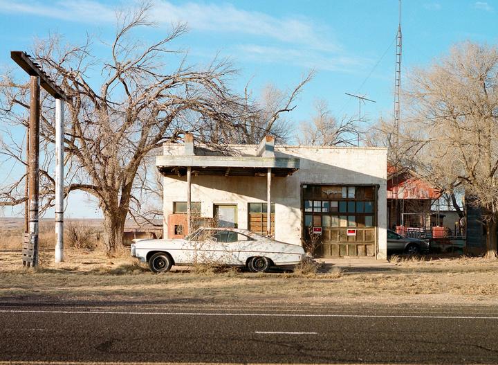 An abandoned white car is parked in front of a dilapidated building with a garage door and a covered entrance.
