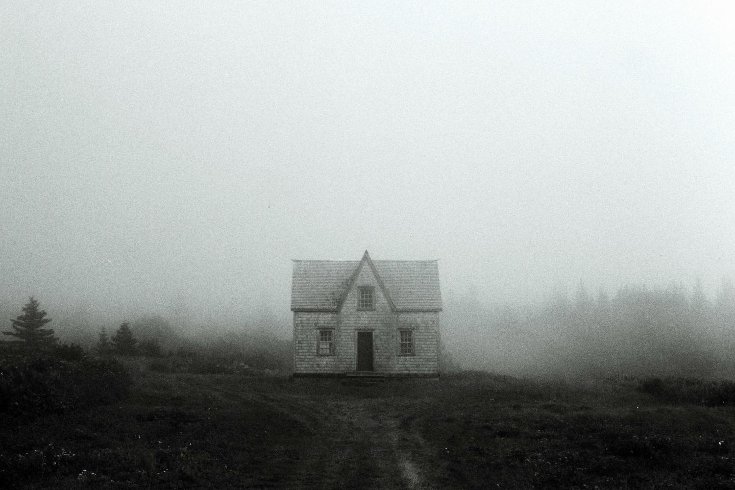 A small house with a peaked roof stands alone in a field on a foggy day.