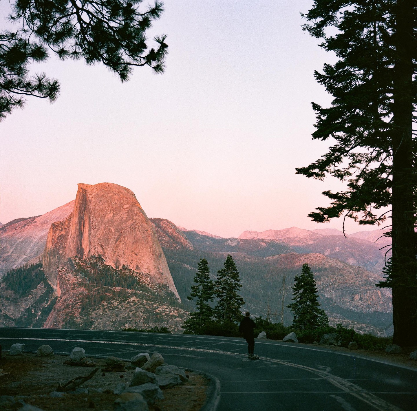 A person on a skateboard cruises down a road with Half Dome and mountains in the background at sunset.