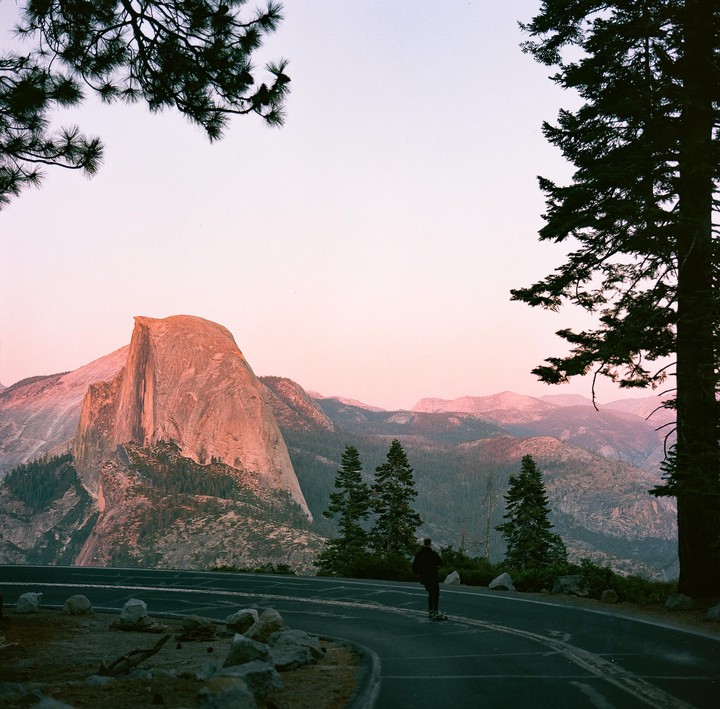 A person on a skateboard cruises down a road with Half Dome and mountains in the background at sunset.