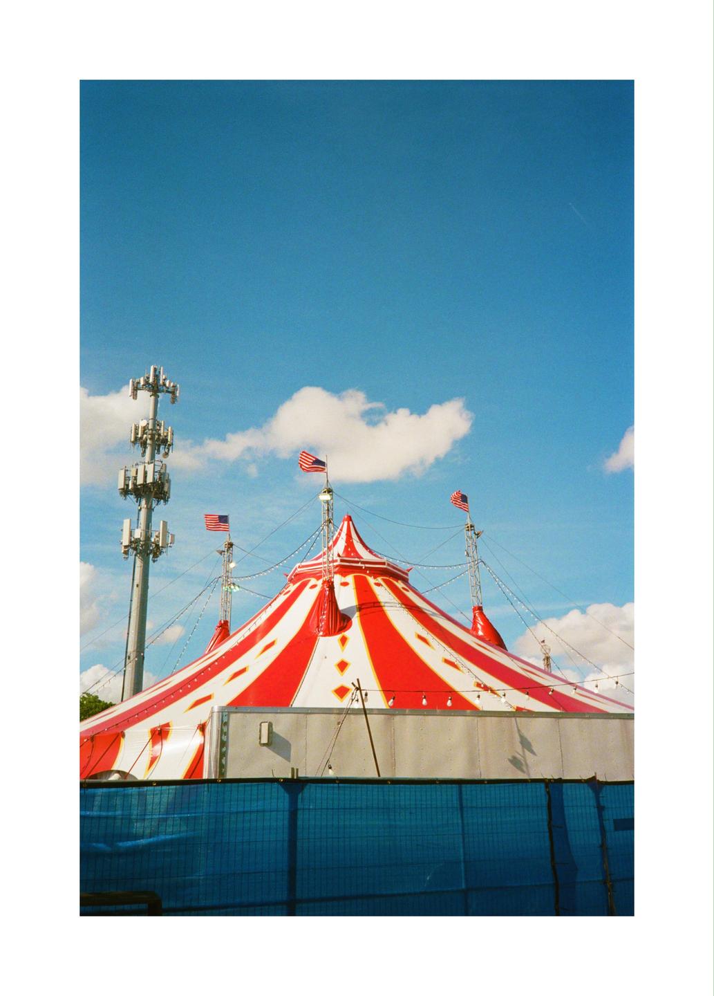 A red and white striped circus tent with American flags on top stands next to a cell tower under a blue sky with white clouds.