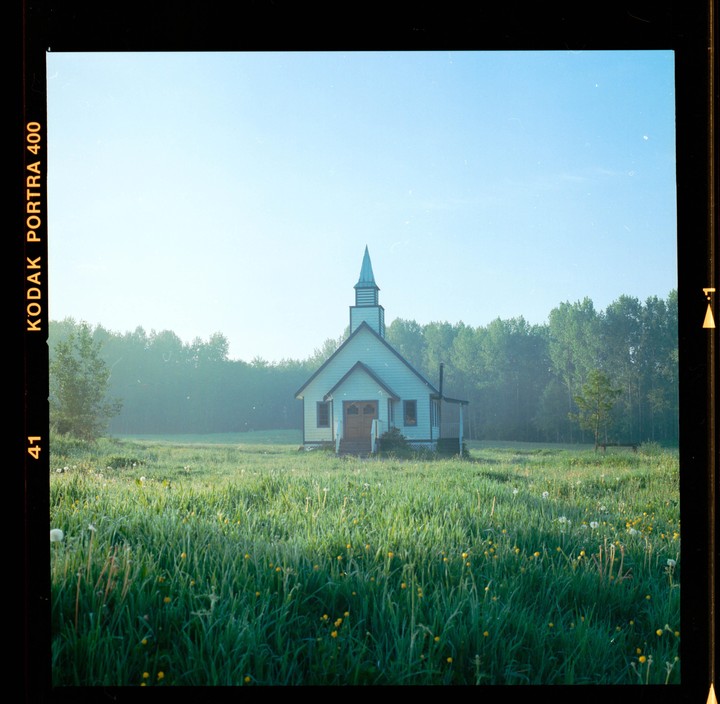 A small white church with a steeple stands in a field of tall grass and wildflowers, with a forest in the background.