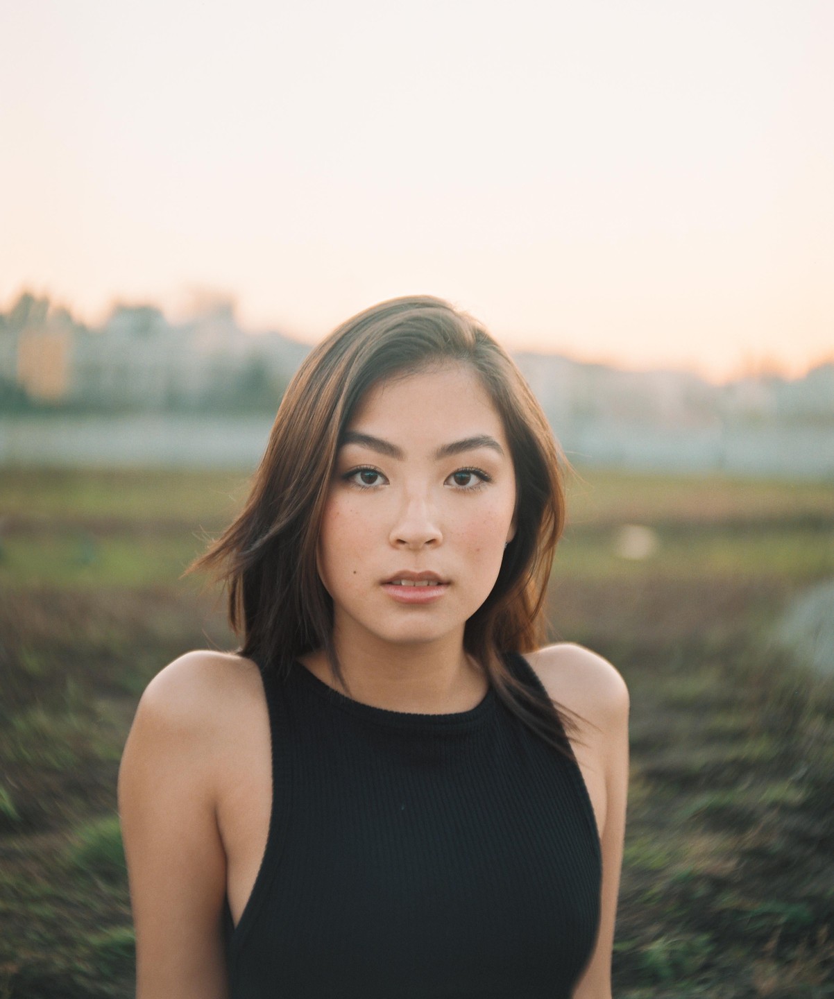 A woman with dark hair looks directly at the viewer with a soft, out-of-focus background of a field and distant buildings.
