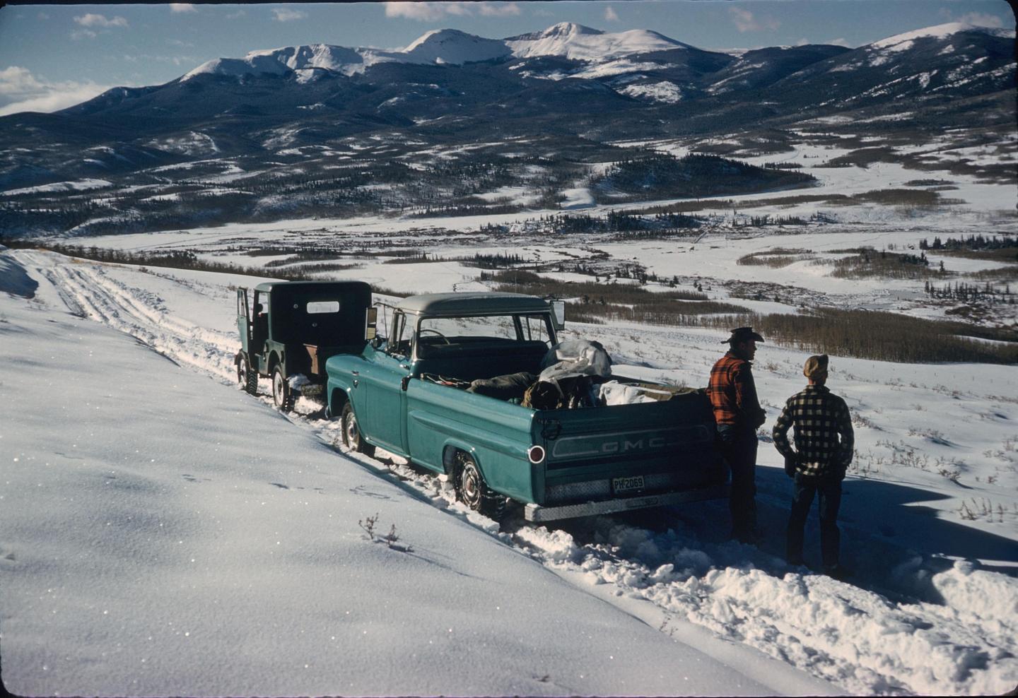 Two men stand next to a pickup truck and a jeep on a snowy mountain road, with snow-capped mountains in the background.