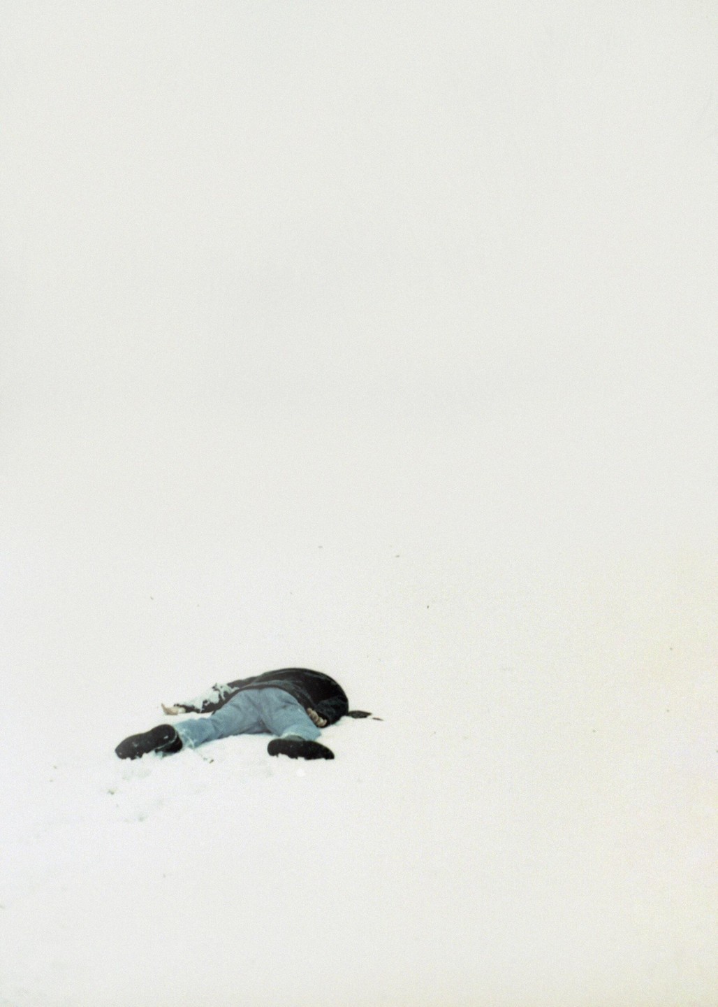 A person lies face down in the snow, wearing a dark jacket and blue jeans.