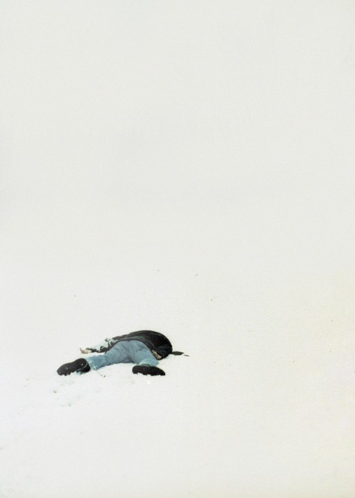 A person lies face down in the snow, wearing a dark jacket and blue jeans.