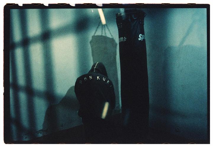 A person in a hooded sweatshirt stands with their back to the viewer next to a punching bag in a dimly lit room.