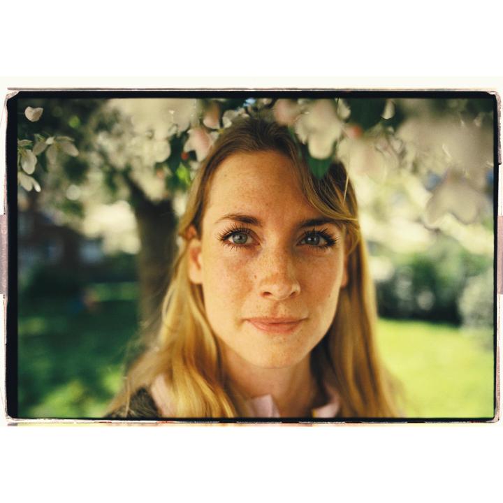 A woman with long blonde hair and freckles looks directly at the viewer, framed by white blossoms.