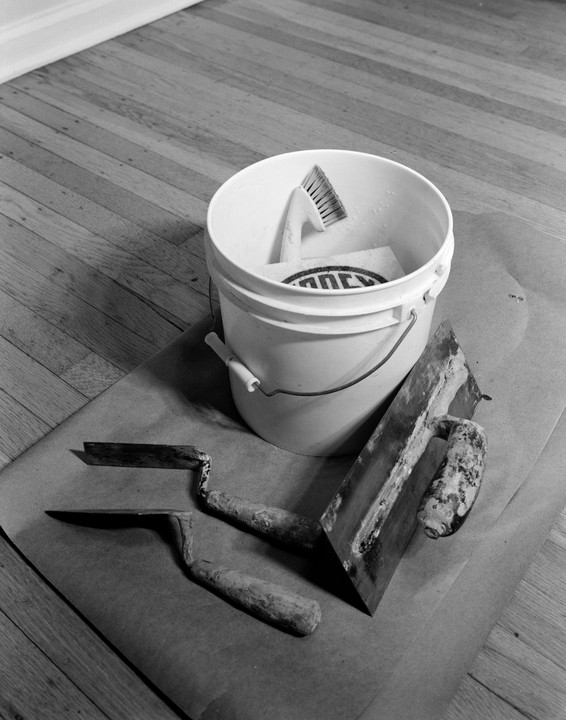 A white bucket filled with tools sits on a brown paper sheet next to two trowels and a float on a wooden floor.