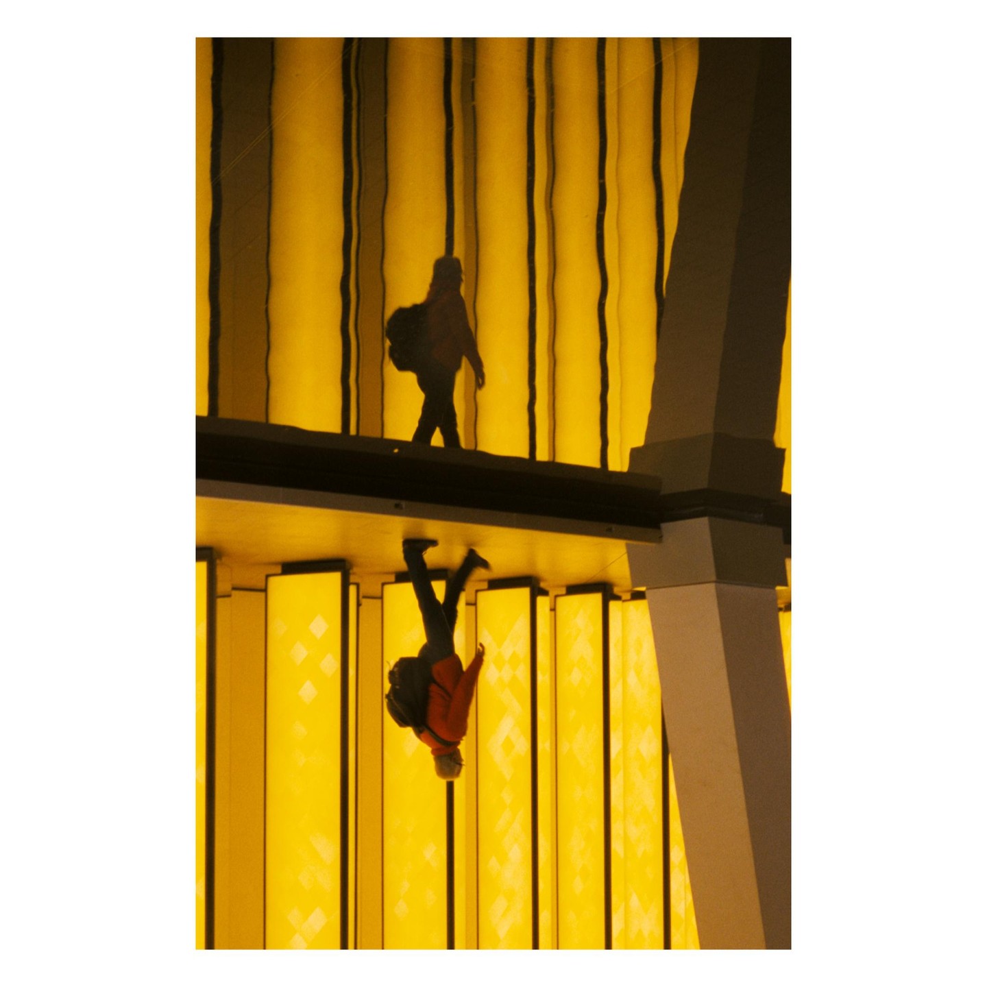 A person walks on a platform above their reflection, both silhouetted against a bright yellow background.