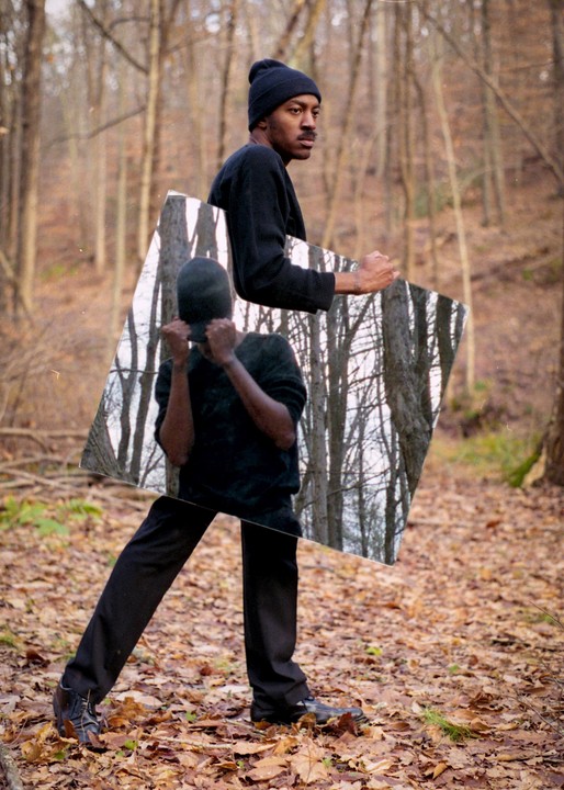A man in a forest holds a mirror that reflects the trees and his own obscured image.