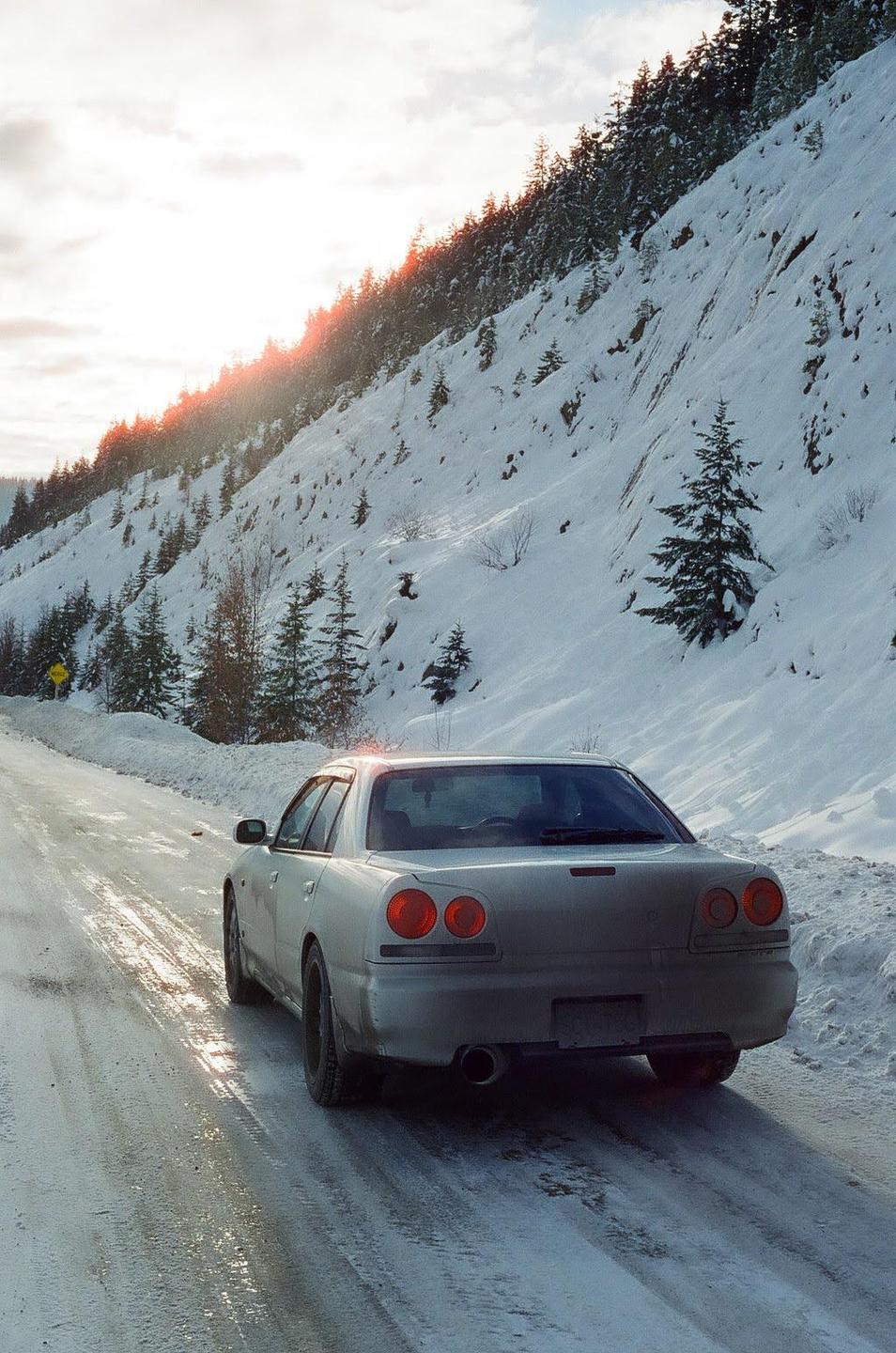 A silver car drives on a snowy road next to a snow-covered mountain with evergreen trees.