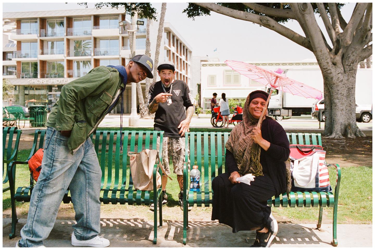 Two men and a woman with a pink parasol pose for a portrait on a green bench in front of a hotel.