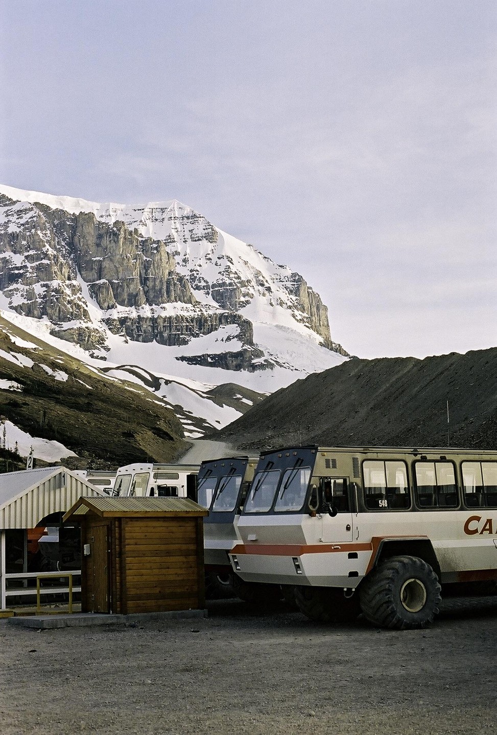 Several large snow buses are parked in front of a snow-covered mountain.
