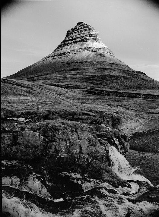 A waterfall cascades over rocks in the foreground with a large, conical mountain rising in the background.