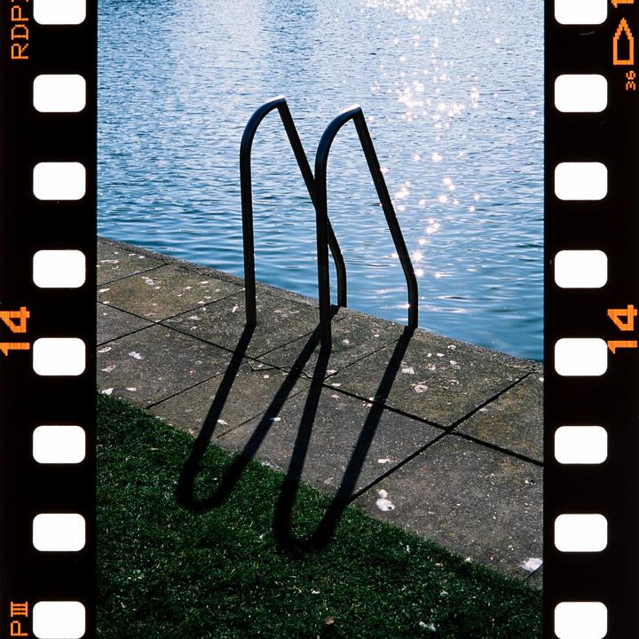 A metal ladder casts long shadows on a concrete edge next to sparkling water.