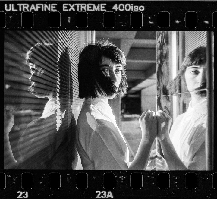 A woman with short hair stands next to a window with blinds, her reflection visible in a mirror.