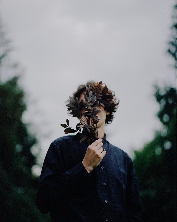 A man with curly hair holds a branch with dark leaves in front of his face, obscuring it.
