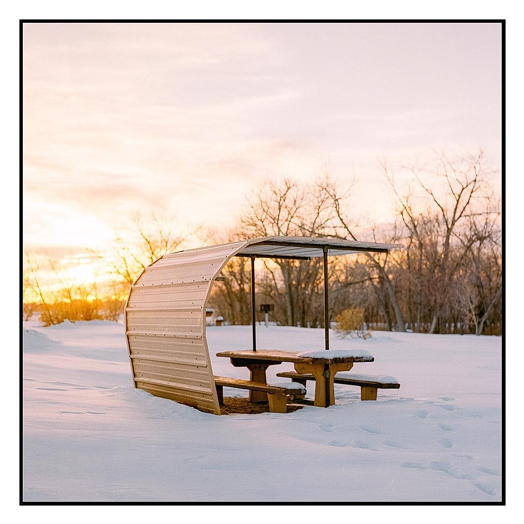 A picnic table with a metal shelter is covered in snow at sunset.