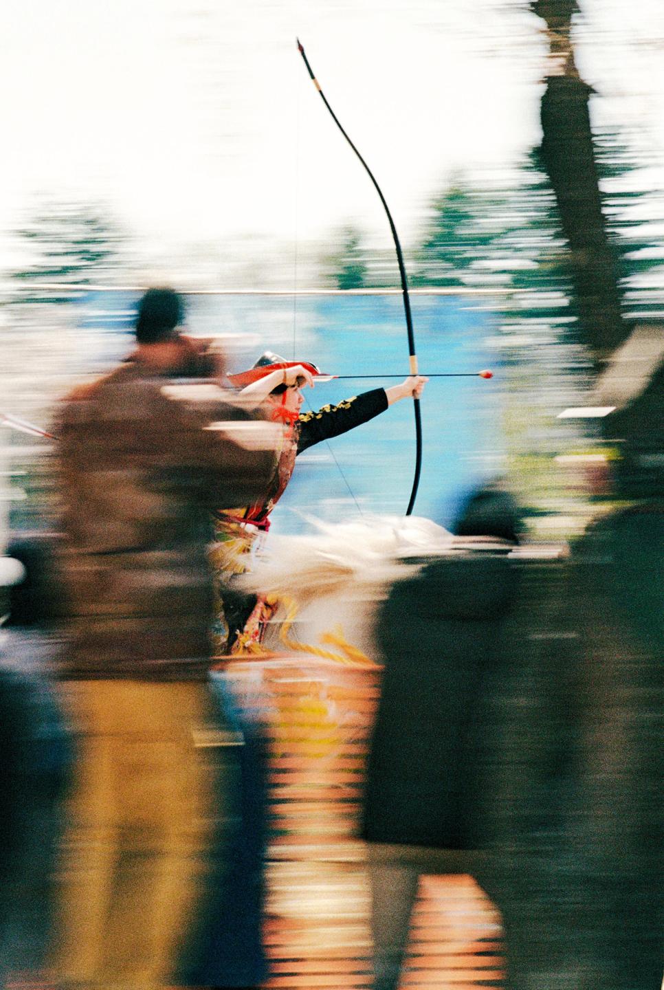A person in traditional attire aims a bow and arrow while riding a horse, surrounded by blurred spectators.