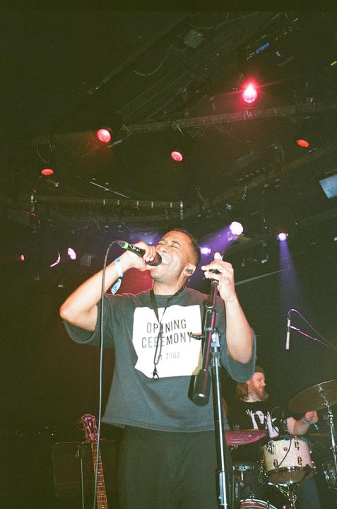 A man sings into a microphone on a stage with a drummer and guitar in the background.