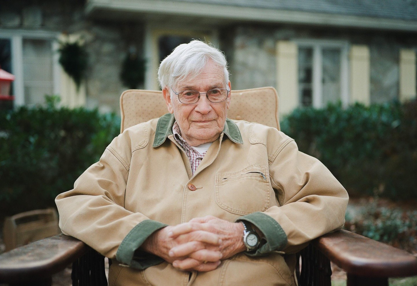 A man with white hair and glasses sits in a chair with his hands clasped in front of a house.