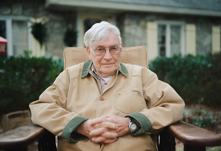 A man with white hair and glasses sits in a chair with his hands clasped in front of a house.