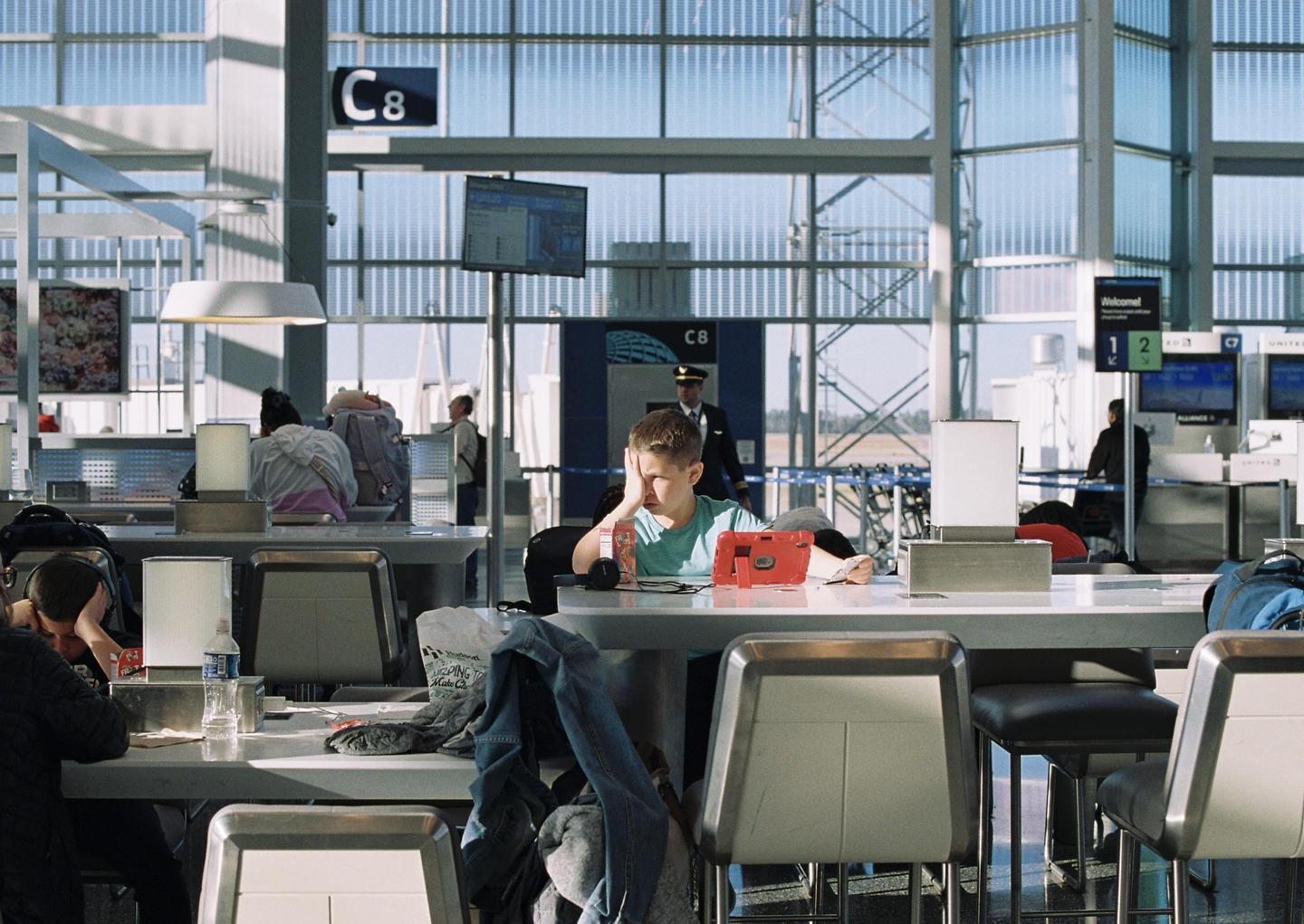 A boy rests his head on his hand while sitting at a table in an airport terminal.