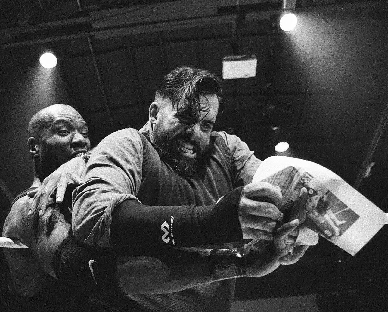 Two men wrestle in a ring, one holding a crumpled newspaper.