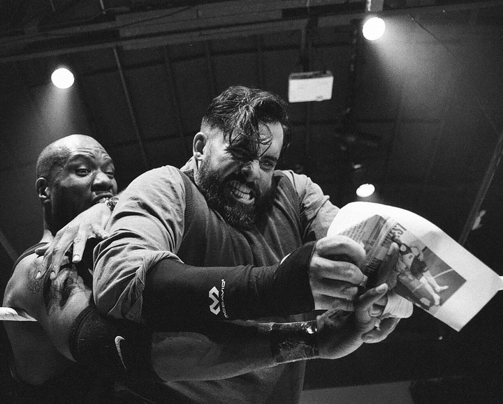 Two men wrestle in a ring, one holding a crumpled newspaper.