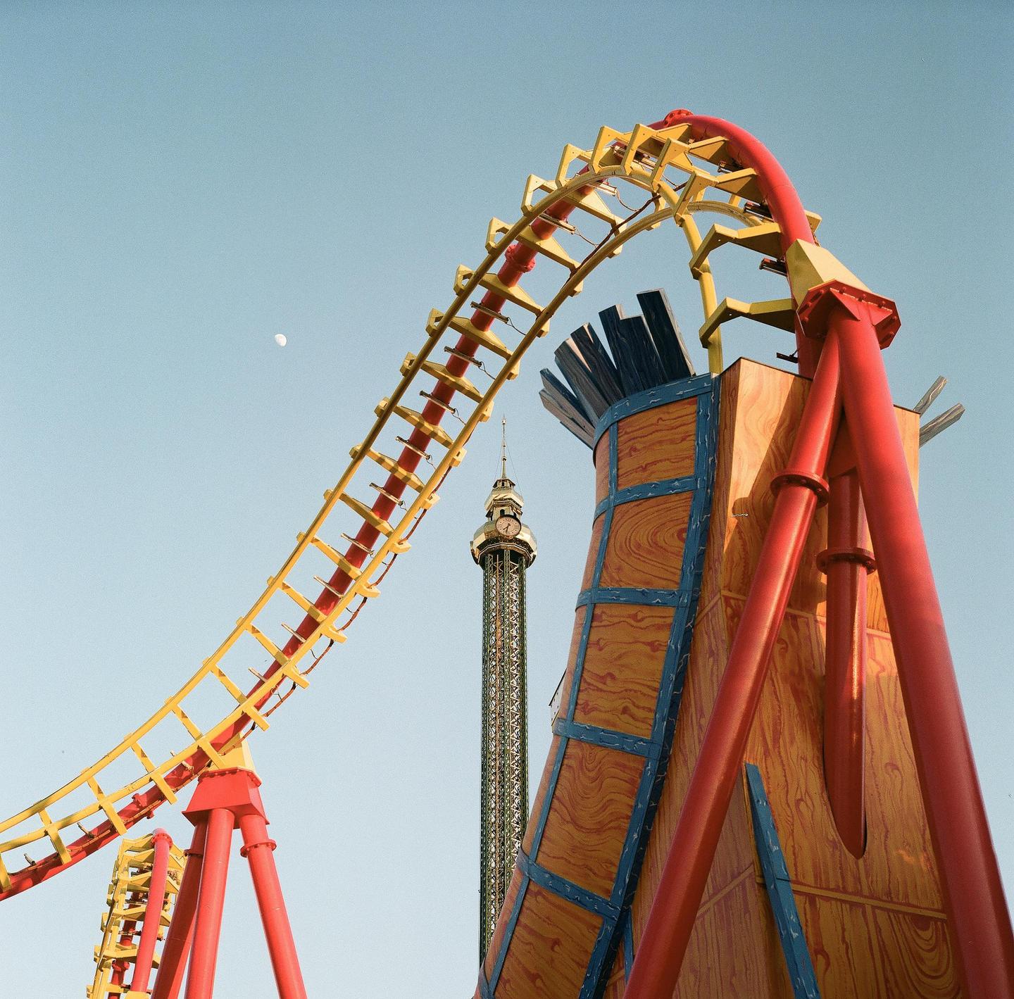 A red and yellow roller coaster track curves against a clear sky with a distant clock tower and a small moon.