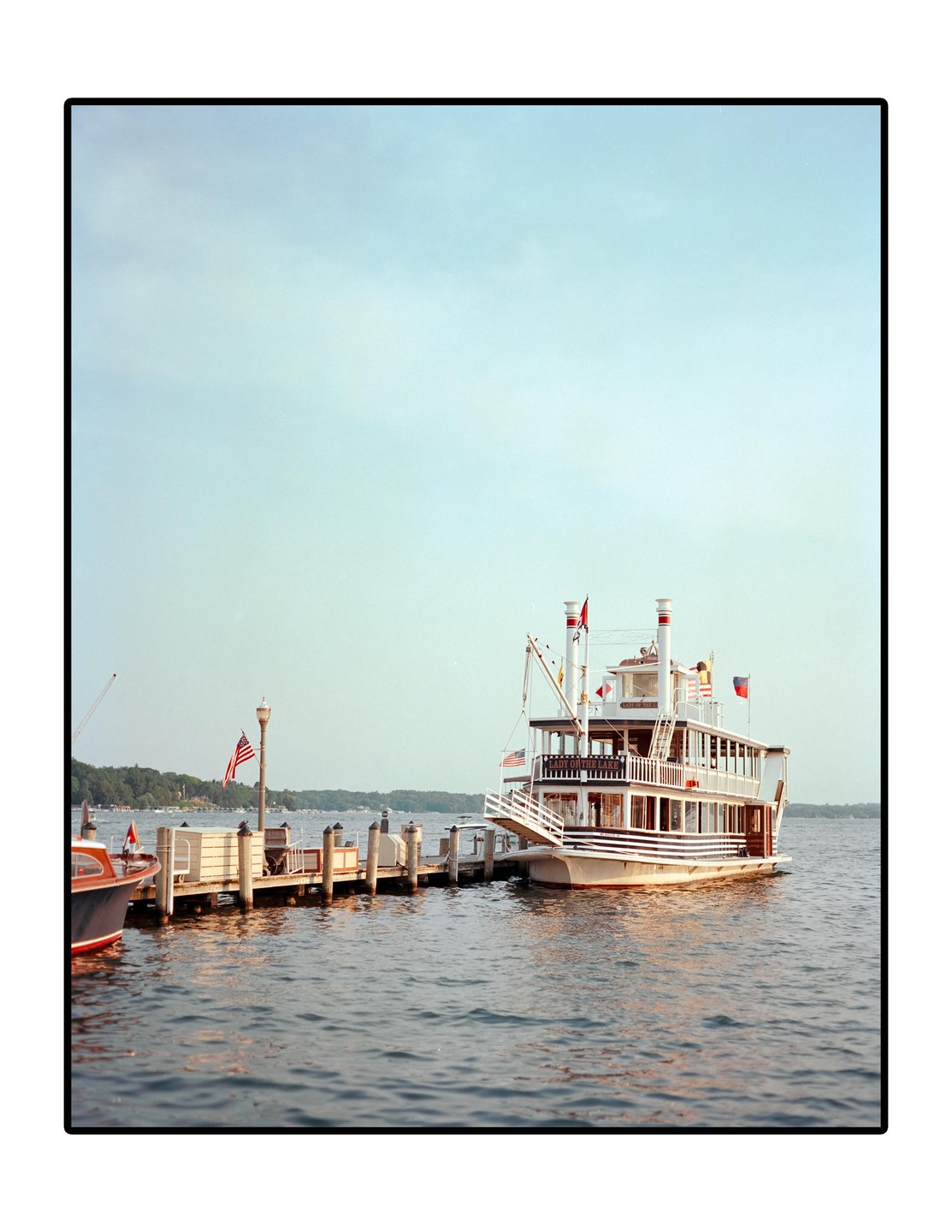 A large white paddle boat is docked at a pier on a lake with trees in the background.