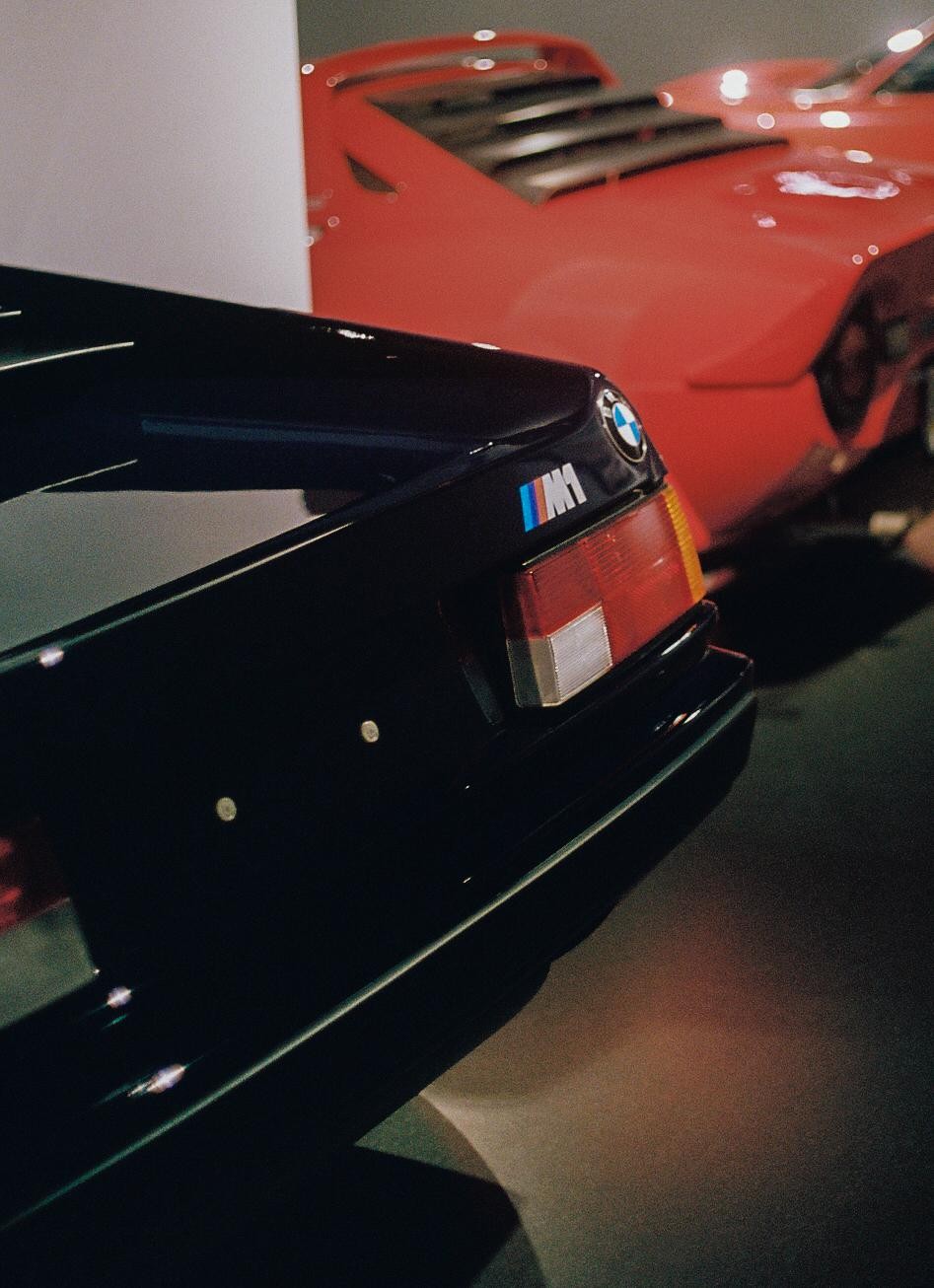 The rear of a black car with a red car in the background.