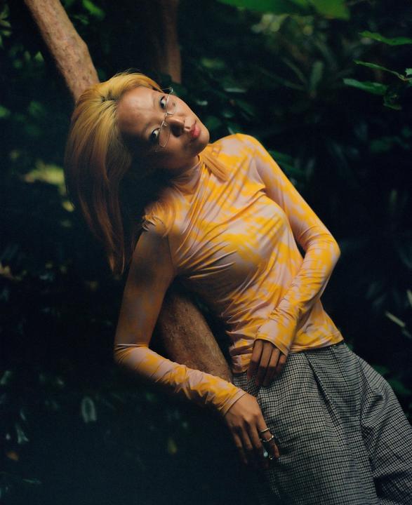 A woman with blonde hair and glasses leans against a tree trunk in a forest.