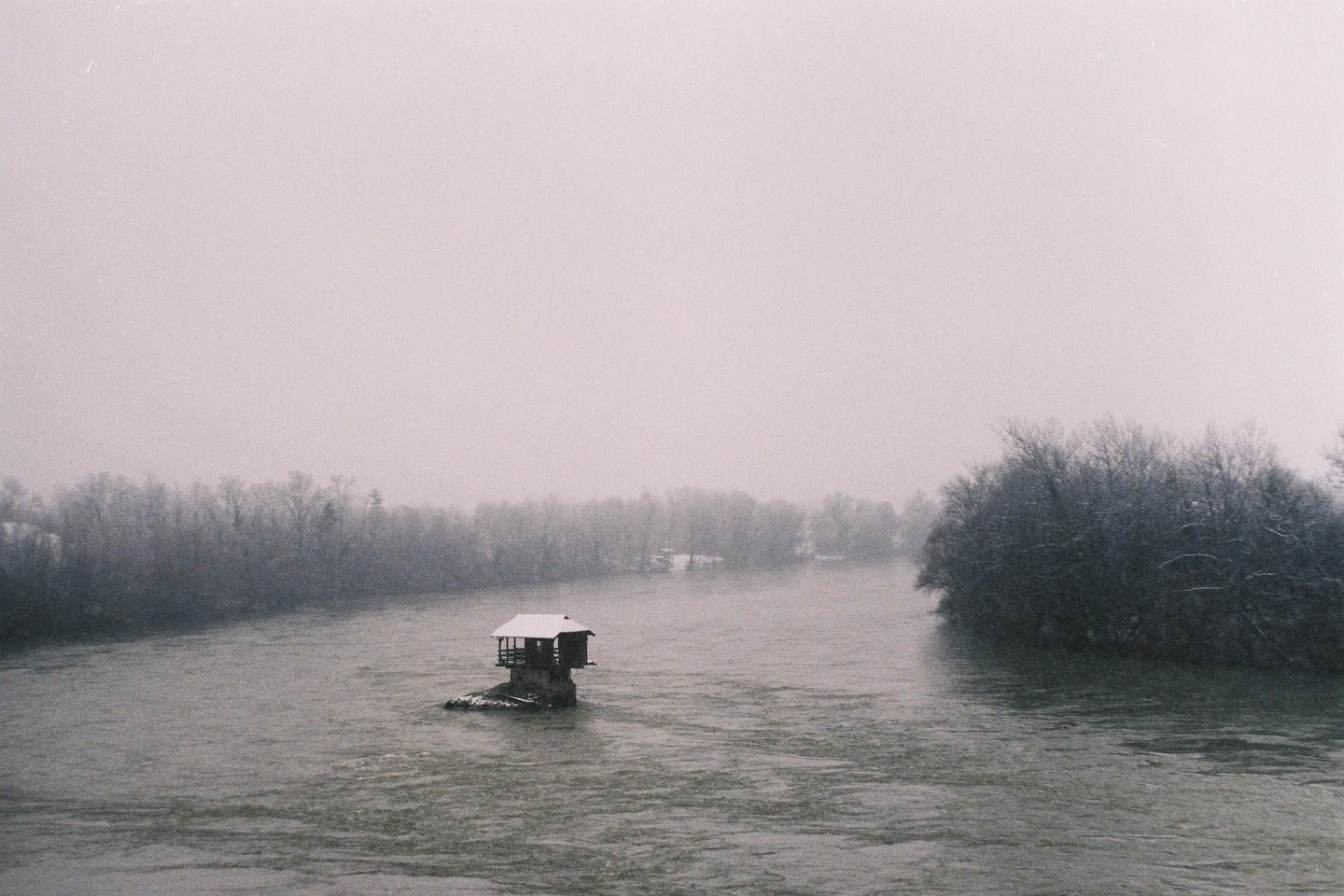 A small house sits on a rock in the middle of a wide river, surrounded by snow-covered trees and a hazy sky.