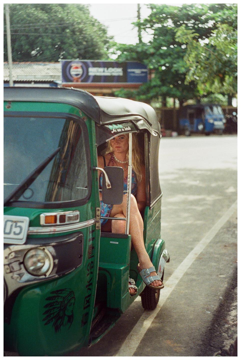A woman with long blonde hair sits in the passenger seat of a green tuk-tuk on a street.