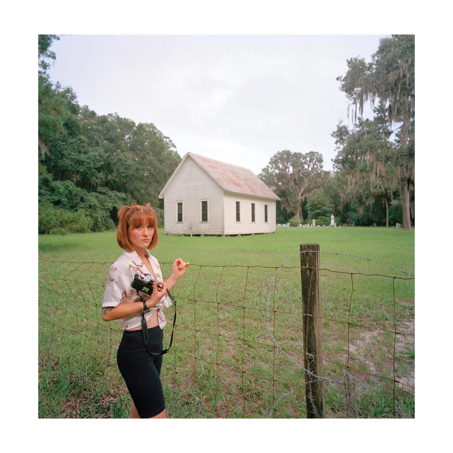 A woman stands in front of a wire fence with a white church and trees in the background.