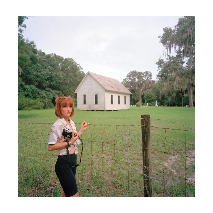 A woman stands in front of a wire fence with a white church and trees in the background.