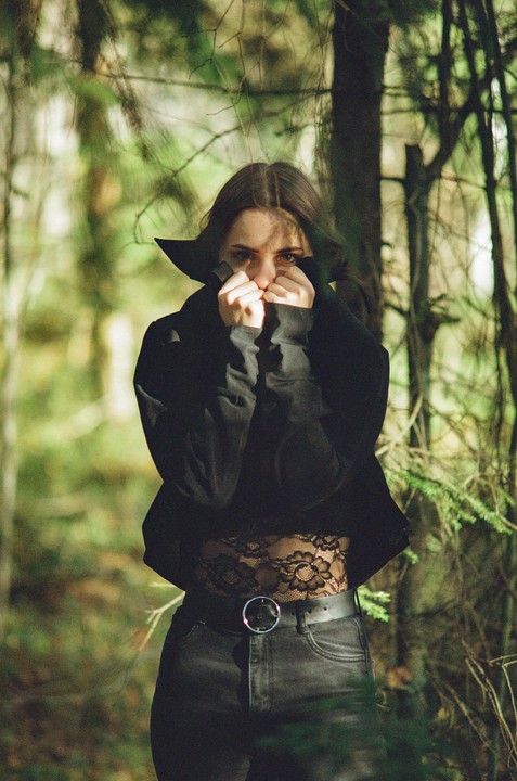 A woman in a black jacket and lace top covers her face with her hands in a forest.