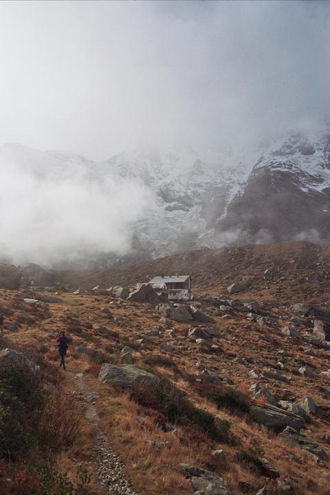 A person walks on a rocky path towards a mountain hut with snow-capped mountains in the background.