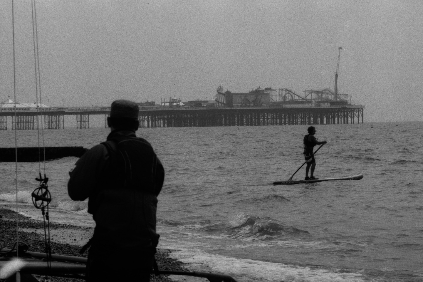 A person stands on a paddleboard in the water with a pier in the background, while another person stands on the shore.