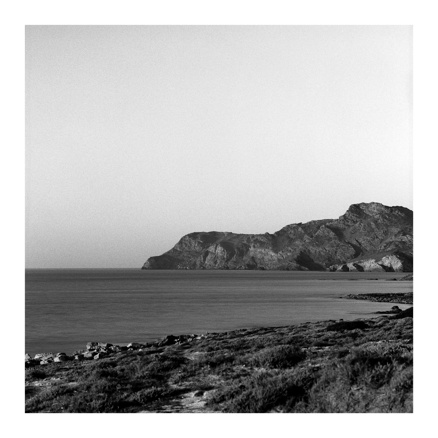 A rocky coastline with a calm sea and a large mountain in the background under a bright sky.