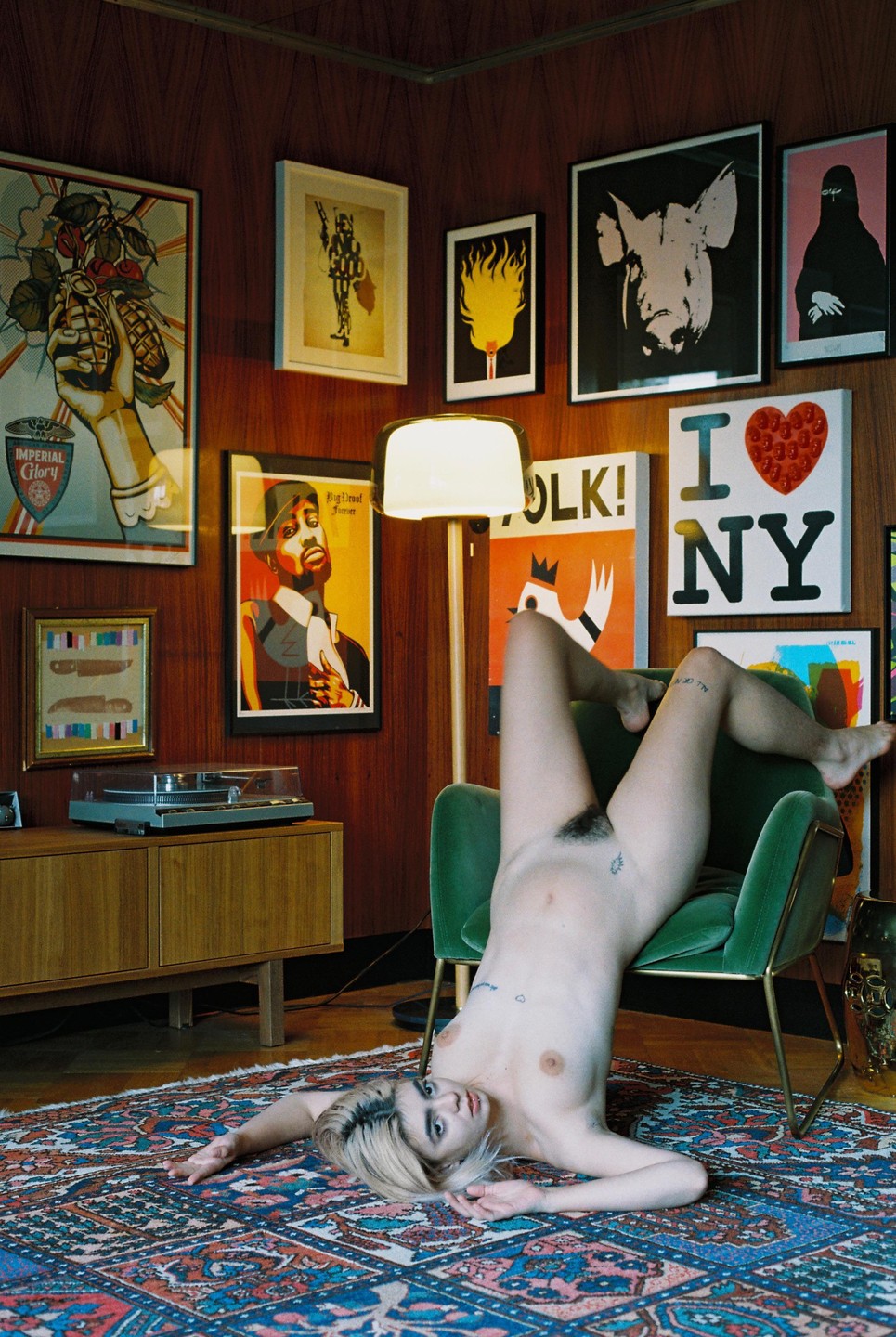 A woman lies on a patterned rug with her legs draped over a green armchair in a room with wood paneling and framed art.