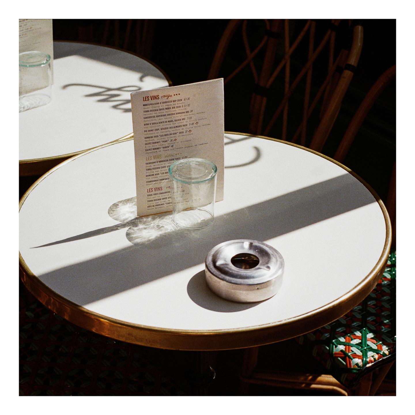 A menu, a glass, and an ashtray sit on a round table in dappled sunlight.